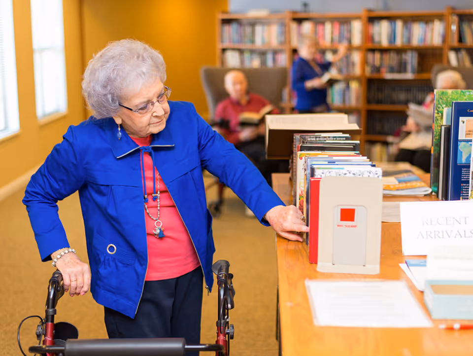 An elderly woman with gray hair, wearing glasses, a blue jacket, and a red shirt, uses a walker while browsing books on a wooden table in a library room. In the background, other elderly individuals are seated and standing, reading books near bookshelves filled with books.