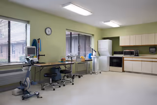 A spacious room with light green walls and white flooring featuring exercise equipment including a stationary bike, a table with stools, and a stacked washer and dryer next to a kitchen area with cabinets, a microwave, and an oven. Two large windows with blinds allow natural light into the room.