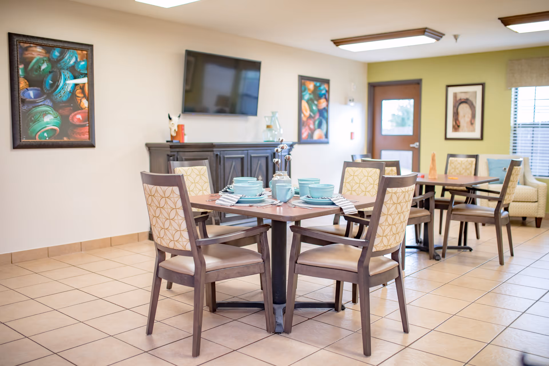 A dining area in a nursing and rehabilitation center with two square wooden tables, each surrounded by four chairs with patterned backs and cushioned seats. The tables are set with turquoise dishes, cups, and napkins. The room has tiled floors, light-colored walls, two framed colorful paintings, a wall-mounted flat-screen TV, a dark wooden cabinet, and a door with a window. There is also a window with blinds and a cushioned armchair in the background.