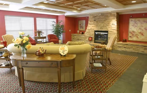 Cozy senior living lounge with sofas and armchairs arranged around a stone fireplace and decorative tables.