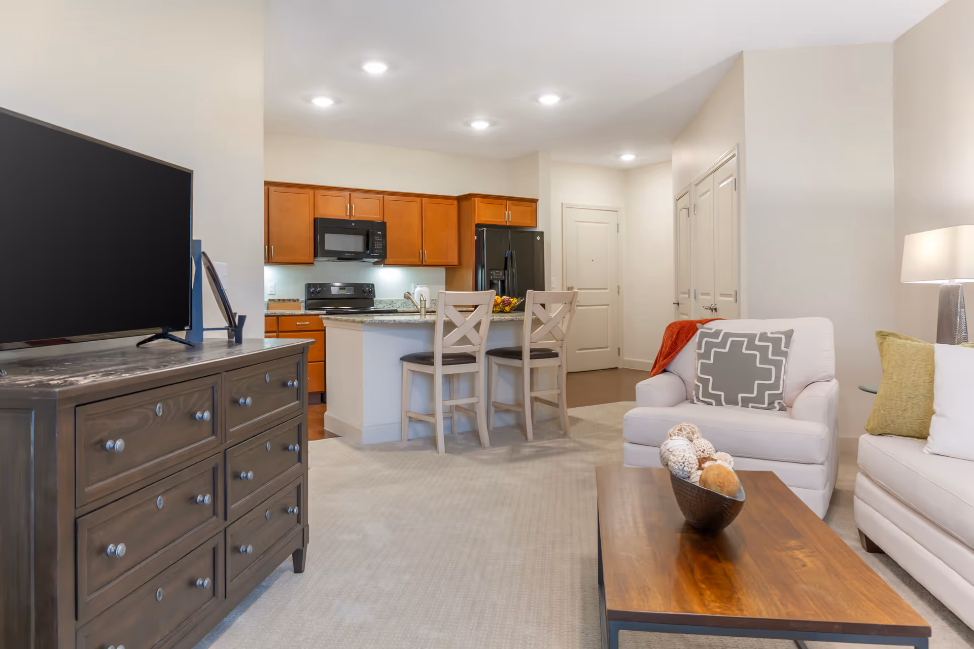 A bright and modern living room and kitchen area in a senior living facility. The living room features a white armchair with a patterned cushion, a white sofa with green and white cushions, a wooden coffee table with decorative items, and a wooden dresser with a flat-screen TV. The kitchen has wooden cabinets, a black microwave, stove, and refrigerator, and a small island with two wooden chairs. The walls are painted light beige, and the floor is carpeted in the living area with hardwood in the kitchen.