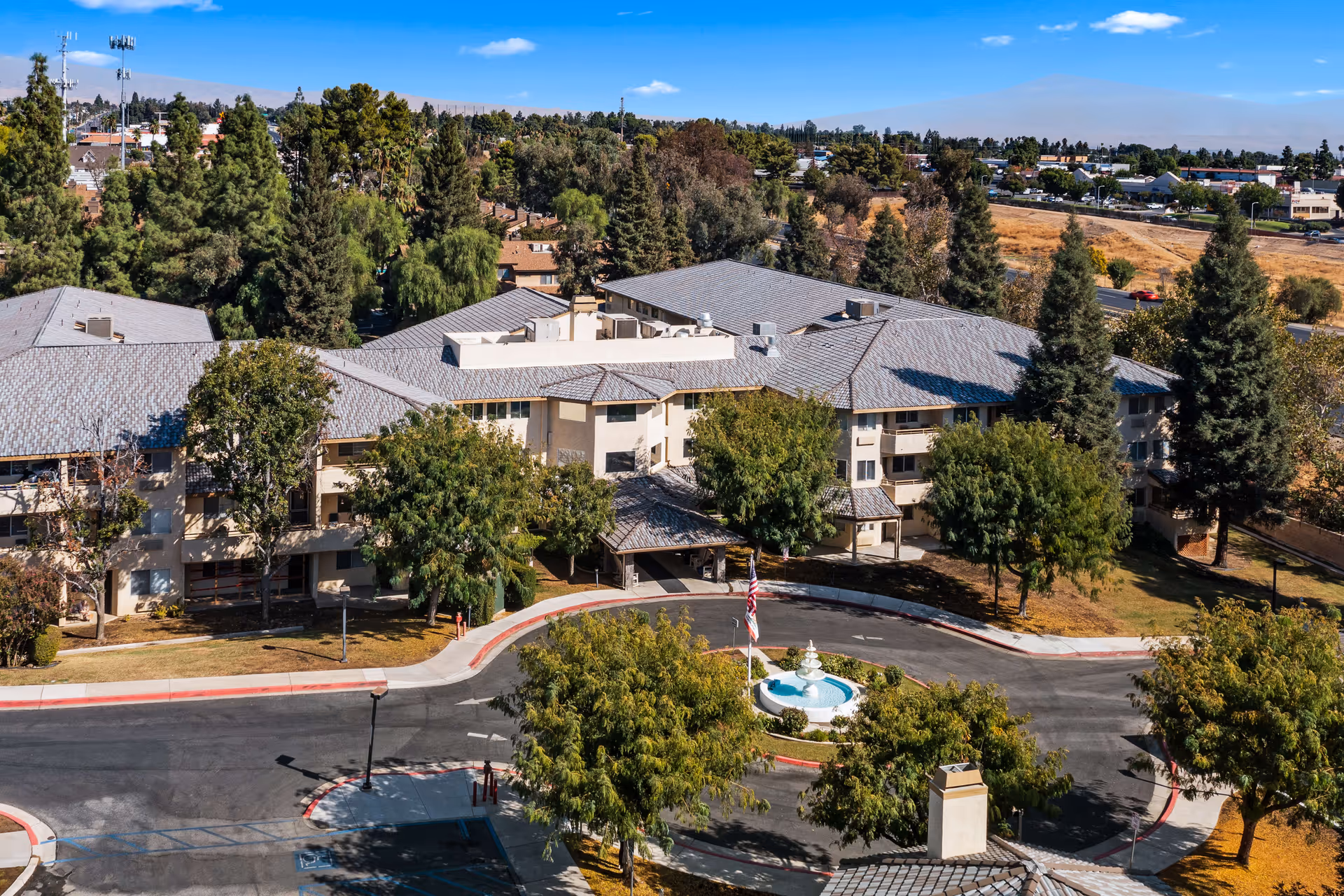 Aerial view of Columbus Estates, a senior living community building surrounded by trees and greenery. The building has a gray roof and beige exterior walls. In front of the building is a circular driveway with a small fountain in the center and an American flag. The surrounding area includes more trees, roads, and other buildings in the distance under a clear blue sky.