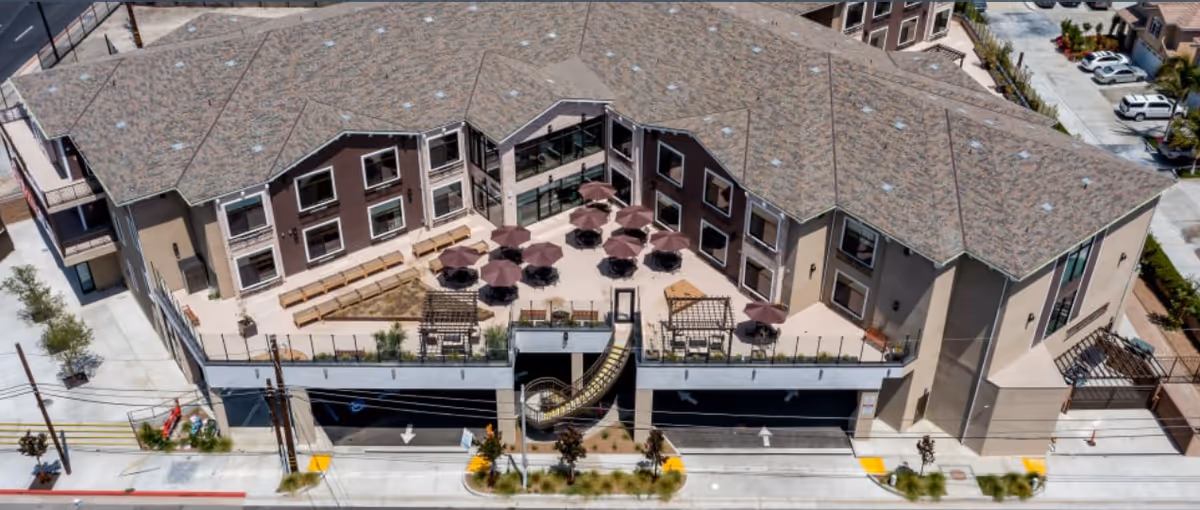Aerial view of a multi-story senior living facility named Beach Terrace, featuring a large outdoor patio area with multiple tables and umbrellas, benches along the walls, and a slide connecting the patio to the ground level. The building has a brown and beige exterior with many windows and a parking area underneath the patio.