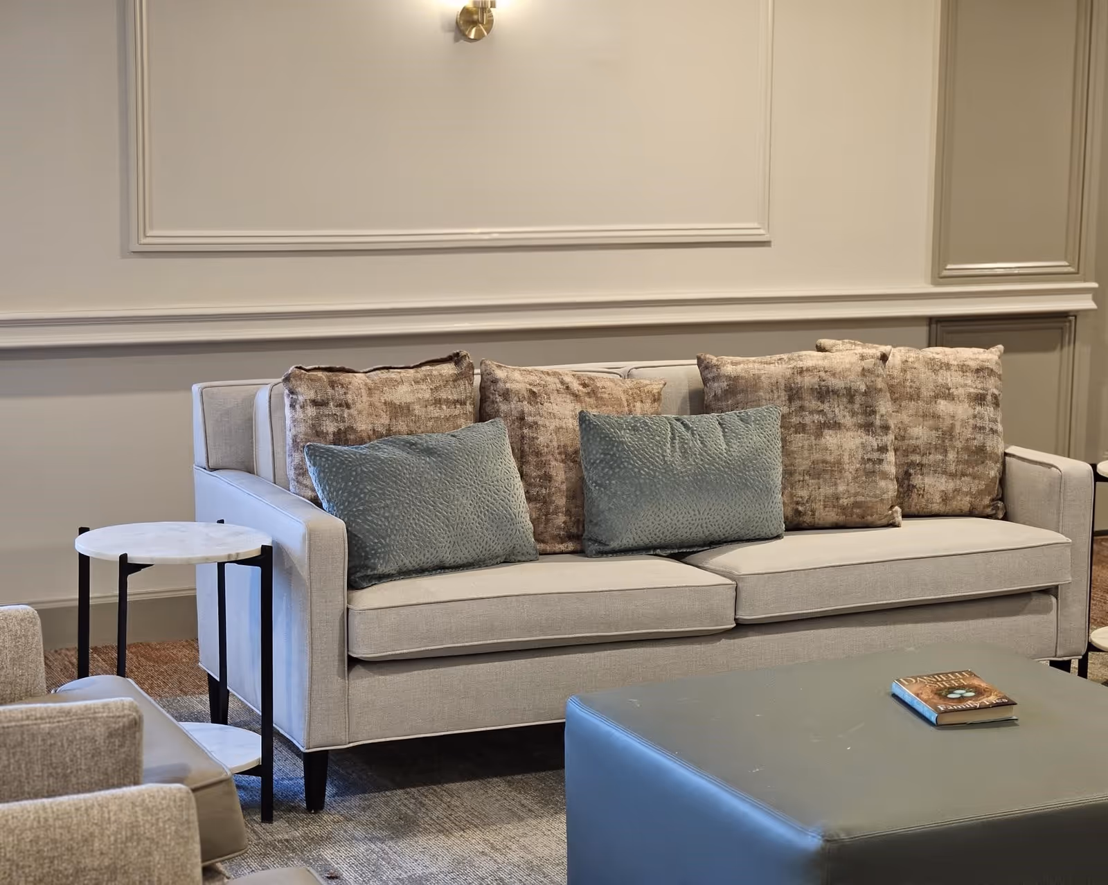 A beige sofa with four brown patterned cushions and two blue textured cushions in a living room setting. Next to the sofa is a small round side table with a marble top and black legs. In front of the sofa is a large blue ottoman with a book placed on it. The wall behind the sofa has decorative molding and a brass wall sconce.