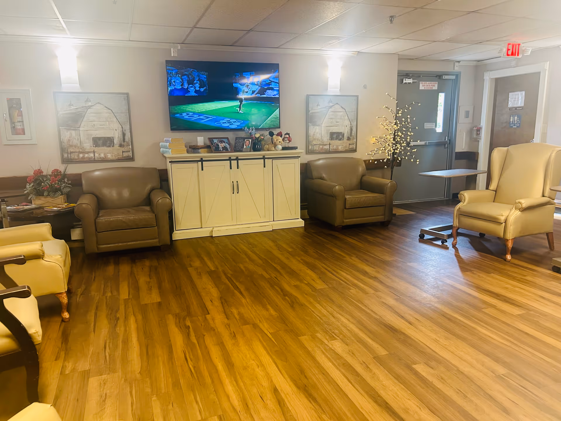 A cozy common area with wooden flooring featuring several armchairs arranged around a white cabinet with a flat-screen TV mounted above it. The walls have two paintings of barns, and there is a decorative lighted tree near the exit door. The room is softly lit with wall sconces and natural light from a window near the door.