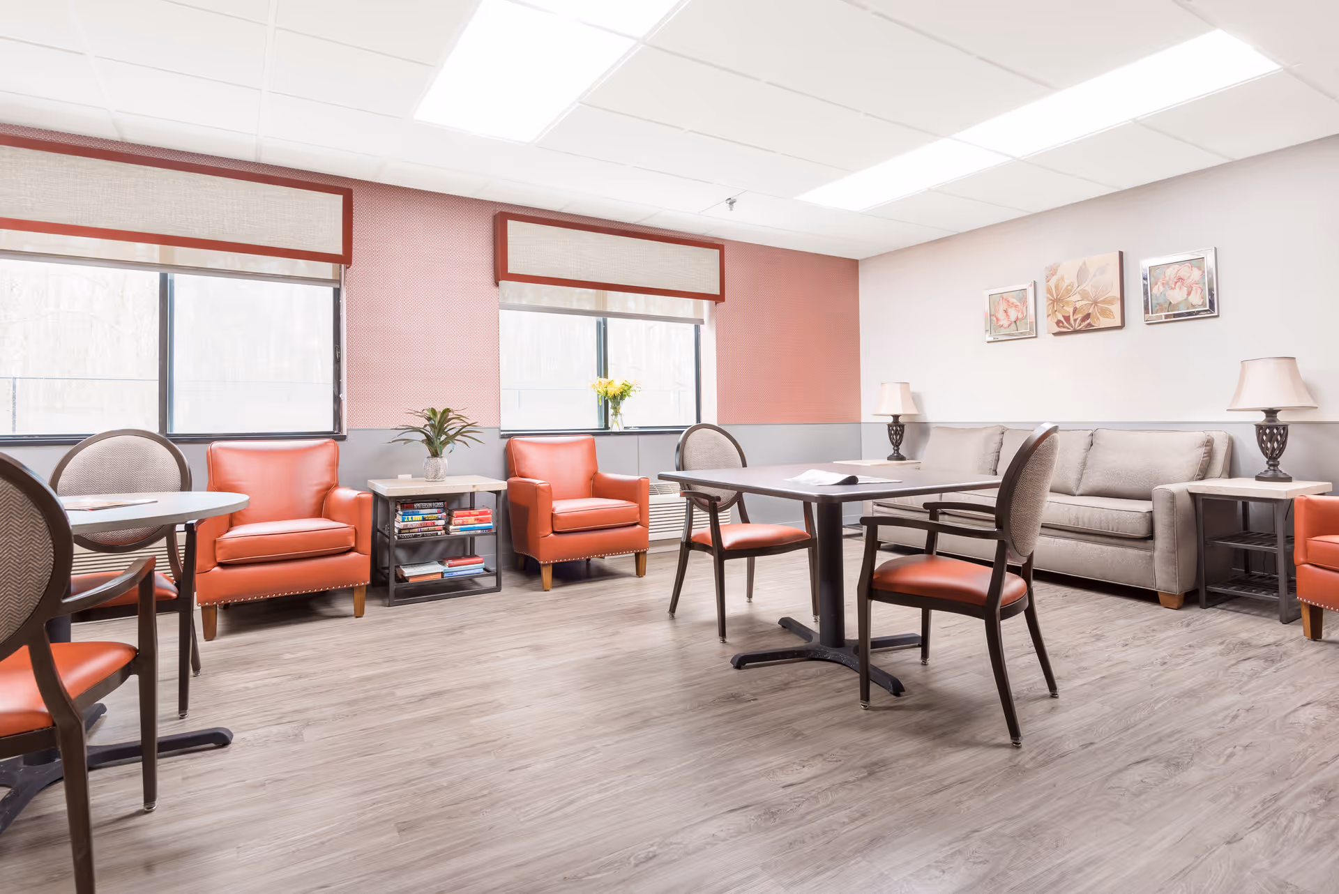 A bright and clean common area in a senior living facility featuring two large windows with beige blinds, two orange armchairs, a beige sofa, two small tables with lamps, two round tables with chairs, and decorative wall art above the sofa. The floor is light wood, and the walls are painted in a combination of light gray and muted red.