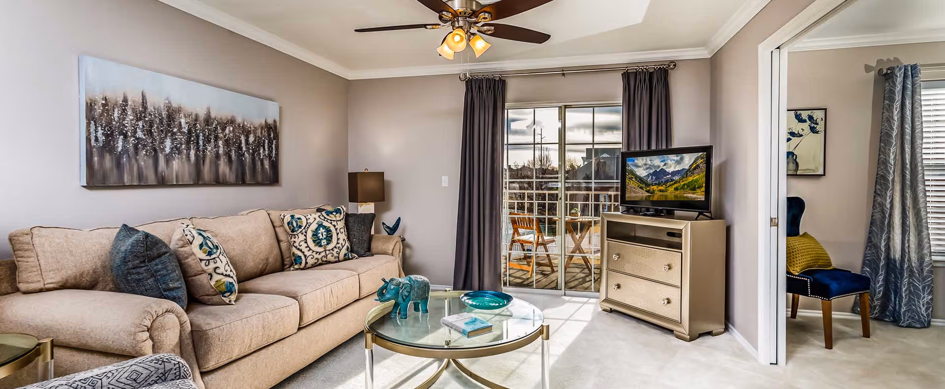 A cozy living room with a beige sofa adorned with patterned and solid pillows, a glass coffee table with decorative items including a small blue elephant figurine and a book, a TV on a beige cabinet showing a mountain landscape, and a ceiling fan with lights. There is a sliding glass door with dark curtains leading to a balcony with a wooden table and chairs. Adjacent to the living room is a small area with a blue chair and a window with curtains.