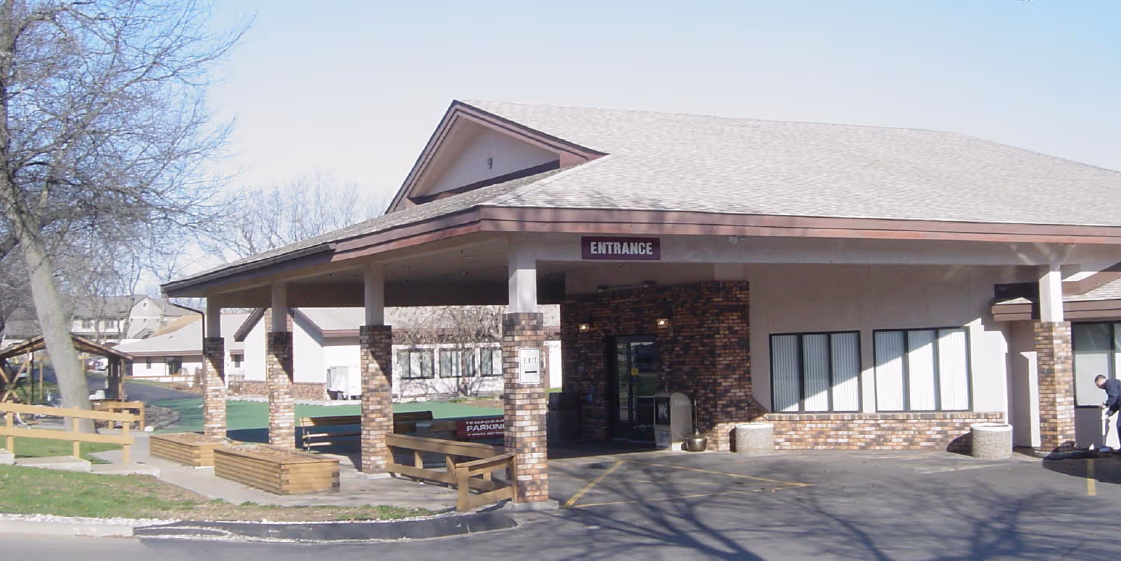 Exterior view of a single-story building with a covered entrance supported by brick columns. The building has a light-colored roof and brick accents around the entrance. There is a sign above the entrance that reads 'ENTRANCE'. Trees and a wooden fence are visible on the left side, and a person is seen near the right edge of the image.