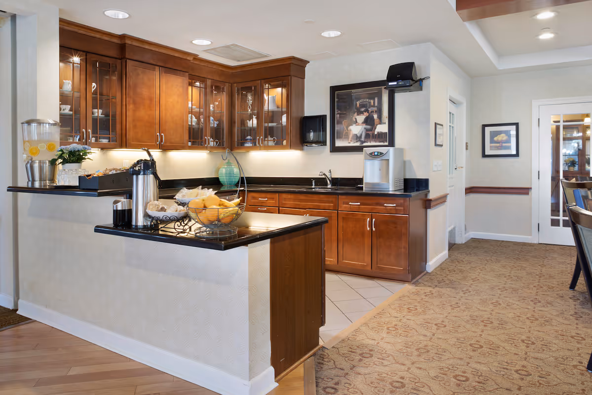 A communal kitchenette with wooden cabinets, black countertops, a sink and a serving counter holding fruit, a coffee urn and beverages.