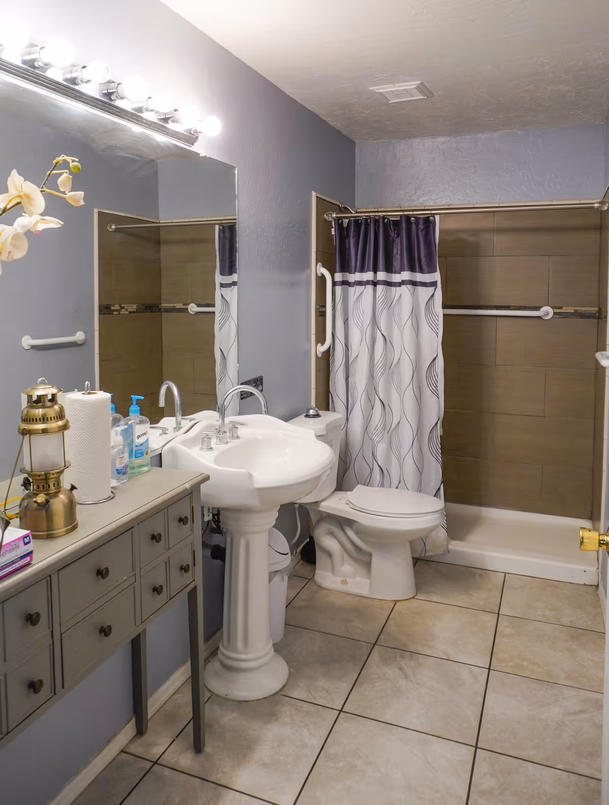 A bathroom with a white pedestal sink, a toilet, and a shower with a curtain featuring a wavy pattern in white and dark purple. The walls are painted gray, and there is a large mirror above the sink with a row of lights. A small table next to the sink holds a vintage lantern, paper towels, hand soap, and other items. The floor is tiled with large beige tiles.