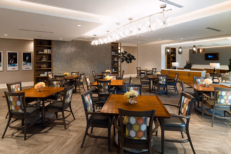 Spacious dining room with wooden tables and patterned chairs, floral centerpieces, bookshelf accents and contemporary overhead lighting.