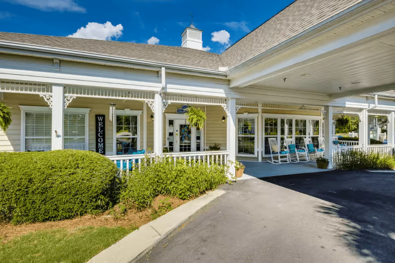Exterior view of The Legacy of Lexington senior living facility showing a covered entrance with white columns and railings, rocking chairs on the porch, hanging plants, and a 'WELCOME' sign near the door under a bright blue sky.