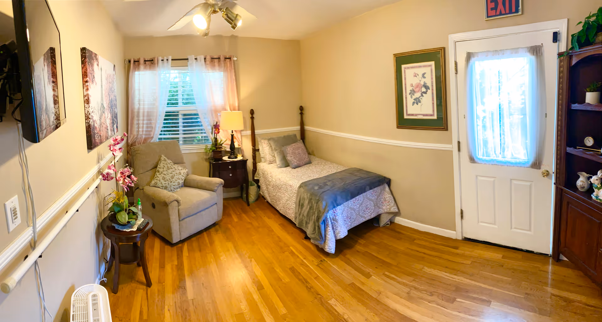 A cozy senior living bedroom with a single bed covered in a patterned quilt and pillows, a beige recliner chair with a decorative pillow, a small round wooden side table with a potted orchid, a window with sheer curtains, a framed floral picture on the wall, a wooden cabinet with decorative items, and a white door with a sheer curtain and an exit sign above it. The room has wooden flooring and soft beige walls with a white chair rail.