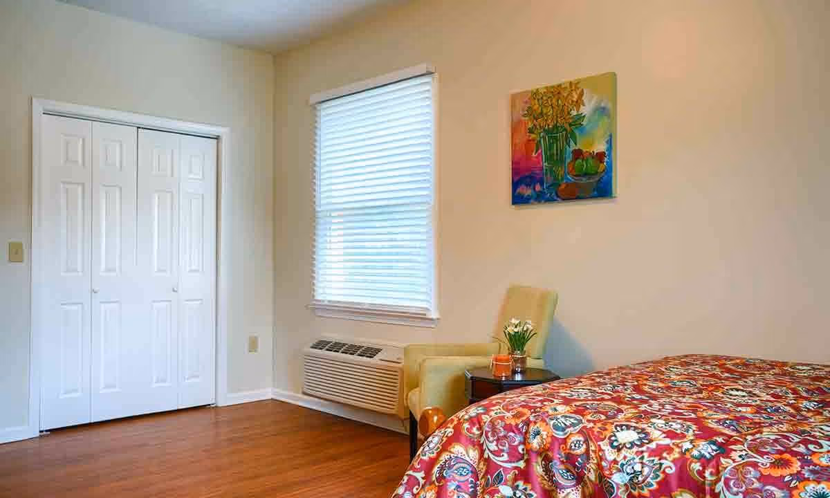A bright bedroom with a colorful patterned bedspread, a beige armchair next to a small dark wooden side table with a small plant and candle, a window with white blinds, a white closet with double doors, and a colorful painting of flowers and fruit on the wall.