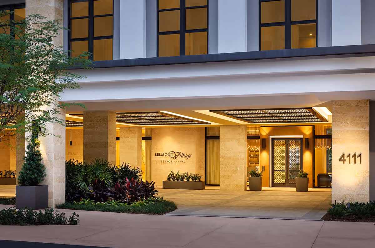 Entrance of Belmont Village Senior Living Coral Gables building with illuminated signage, large windows, stone pillars, decorative plants, and the address number 4111 displayed on the right pillar.