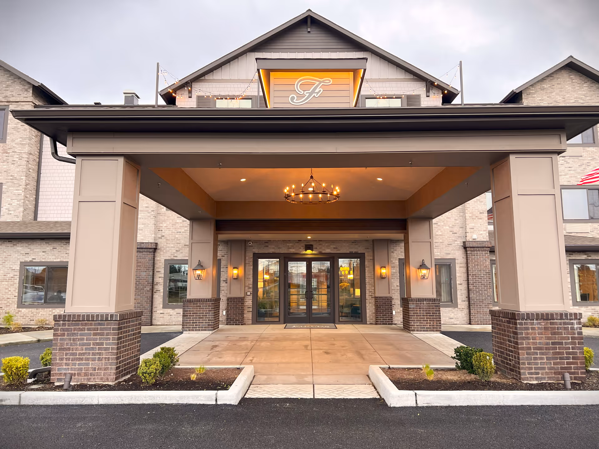 Covered front entrance of a senior living building with a porte-cochere, lit chandelier, glass double doors, and brick-clad columns.
