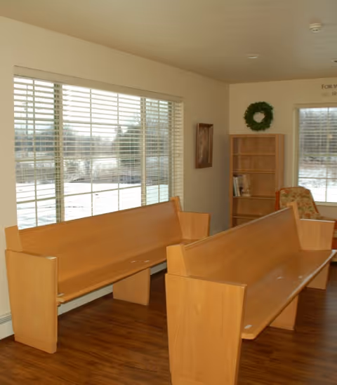 Interior common room with two wooden bench seats by large windows, a small bookshelf and a wreath on the wall.