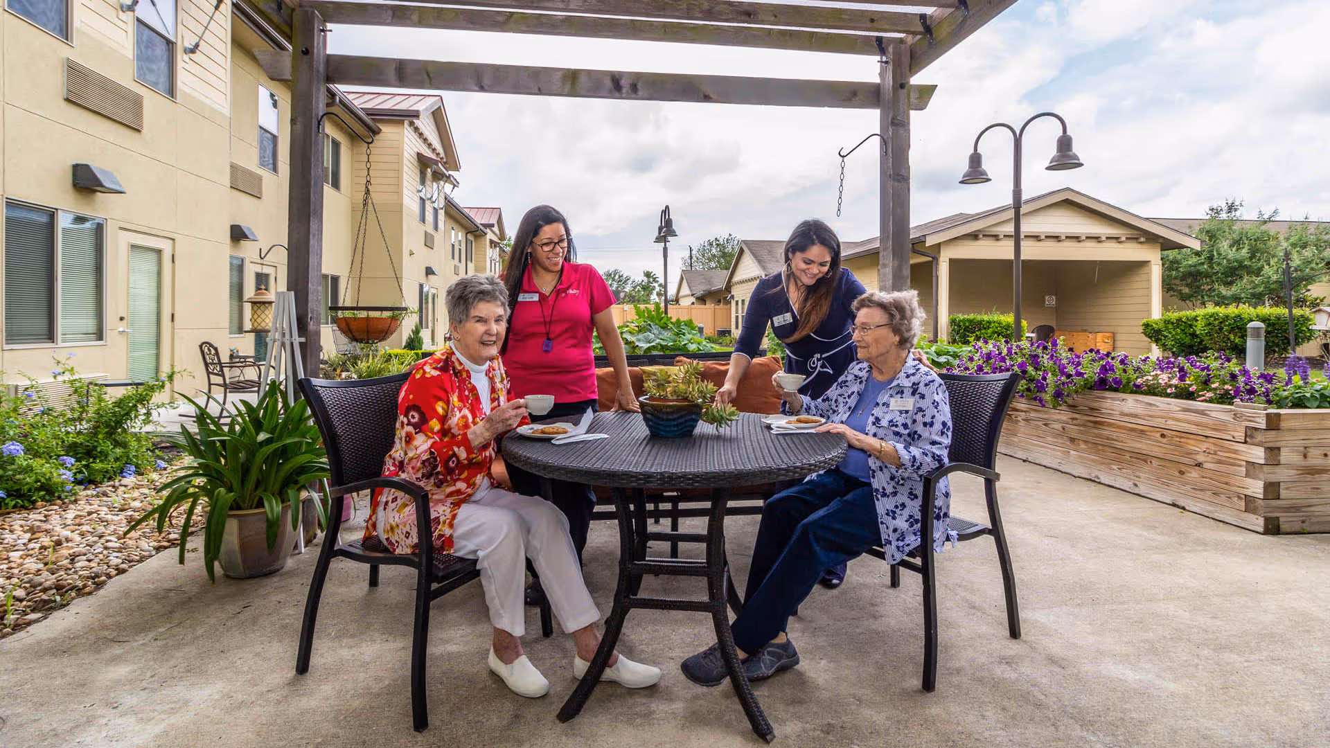 Two elderly women sitting at a round outdoor table under a pergola, enjoying tea and snacks, accompanied by two caregivers standing nearby. The setting is a garden area with flowers, plants, and residential buildings in the background.