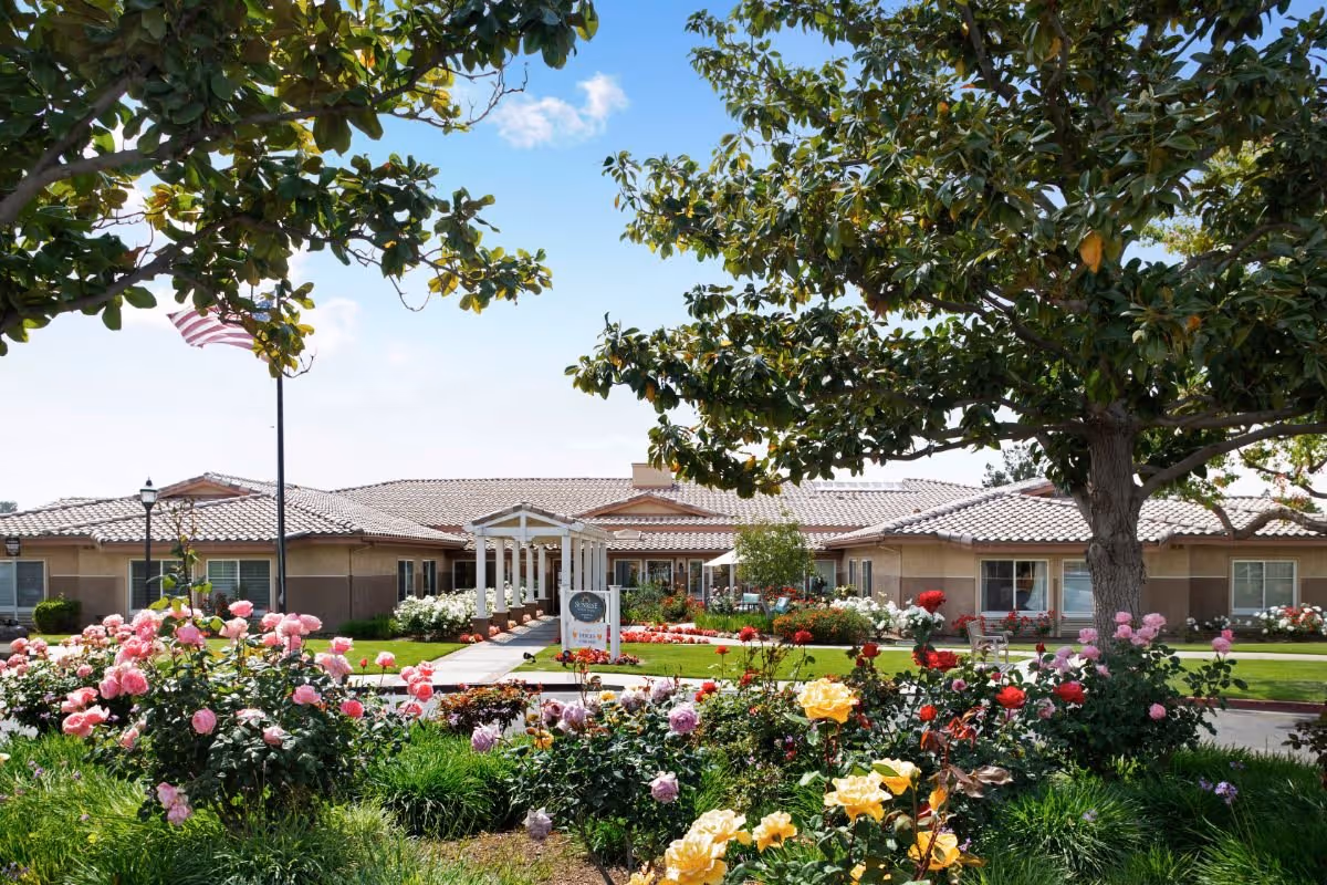 Single-story senior living building with a tiled roof, a white entrance pergola, and a colorful rose garden and lawn in the foreground.
