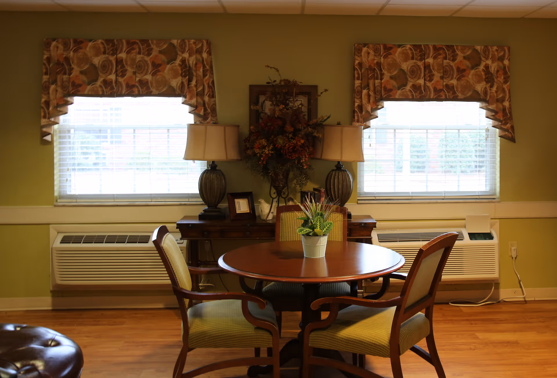 Small dining area with a round wooden table and four chairs in front of two windows, flanked by lamps and a console table with decorative plants and flowers.