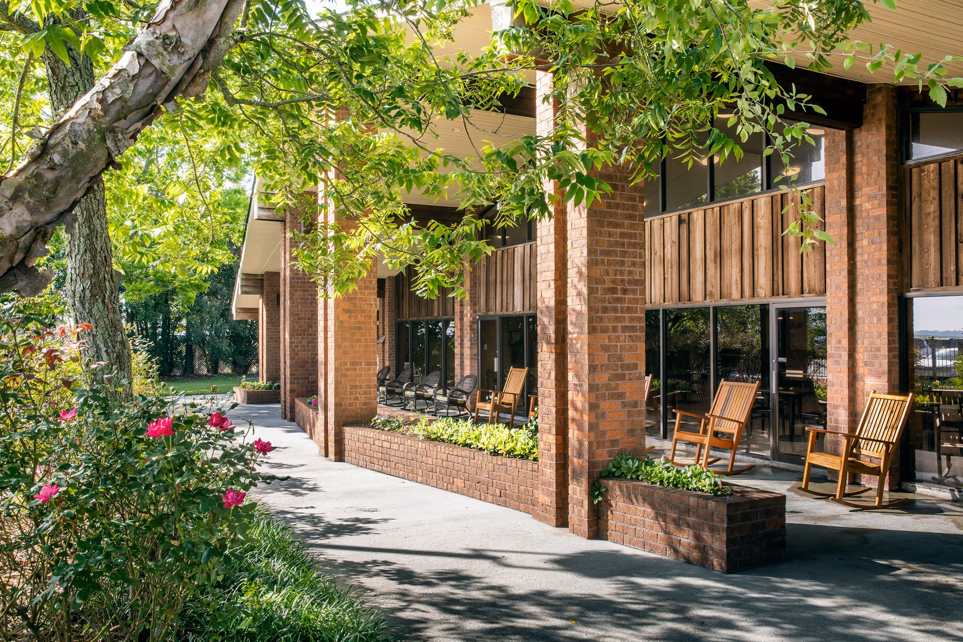 Outdoor patio area of a brick building with wooden rocking chairs, large windows, and green plants along the walkway. Trees and flowering bushes surround the paved path leading to the entrance.