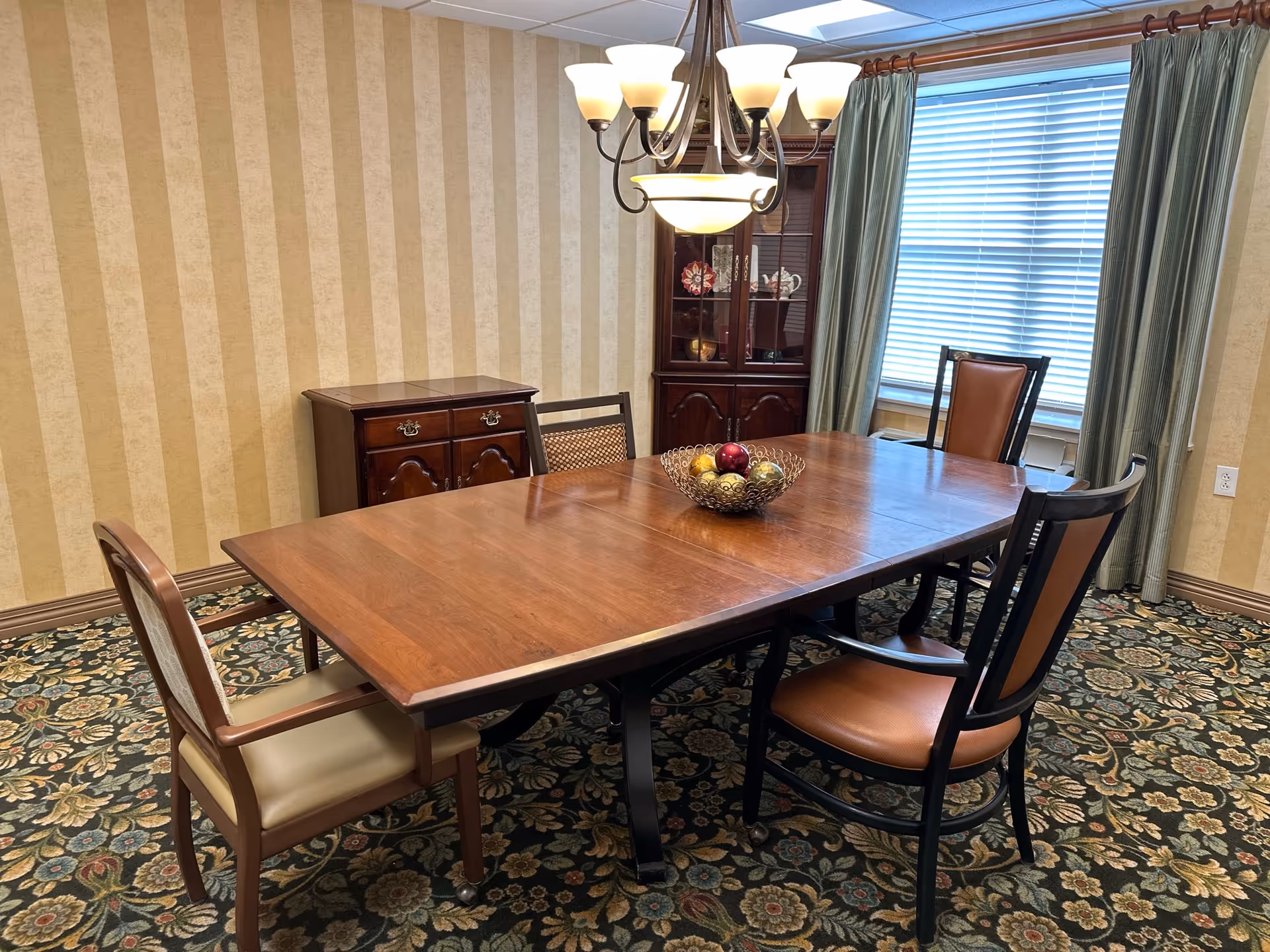 A dining room with a wooden rectangular table surrounded by four chairs with leather seats. A decorative bowl with ornamental balls is placed in the center of the table. The room has patterned carpet flooring, striped wallpaper, a wooden cabinet with glass doors displaying decorative items, a window with closed blinds, and green curtains. A chandelier with multiple lights hangs above the table.