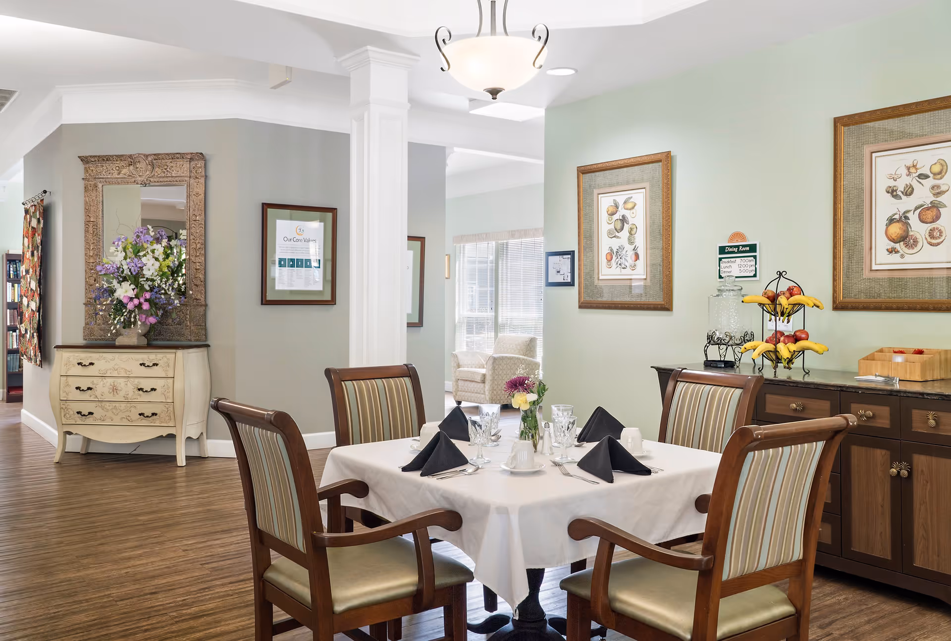 A dining area in a senior living facility with a square table set for four, featuring white tablecloth, black napkins, glassware, and a small flower vase. The room has wooden chairs with striped upholstery, framed botanical prints on the light green walls, and a sideboard with a water dispenser, fruit stand, and wooden box. In the background, there is a decorative chest with a large ornate mirror and a floral arrangement, and a glimpse of a sitting area with armchairs near a window.