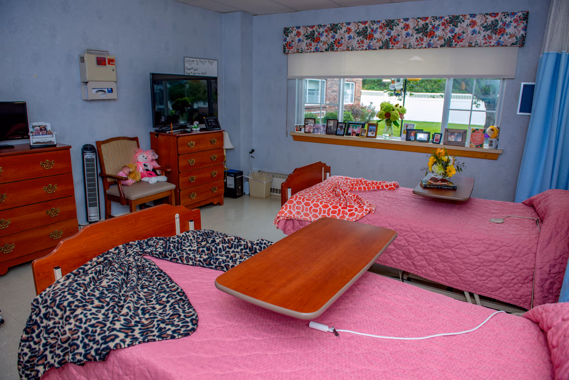 A cozy room with two single beds covered in pink quilts, each with a wooden overbed table. One bed has a leopard print blanket, and the other has a red patterned blanket. There are two wooden dressers, a chair with stuffed animals, a TV, and a window with a floral valance and several framed photos on the windowsill. A vase with flowers is also on one of the beds.