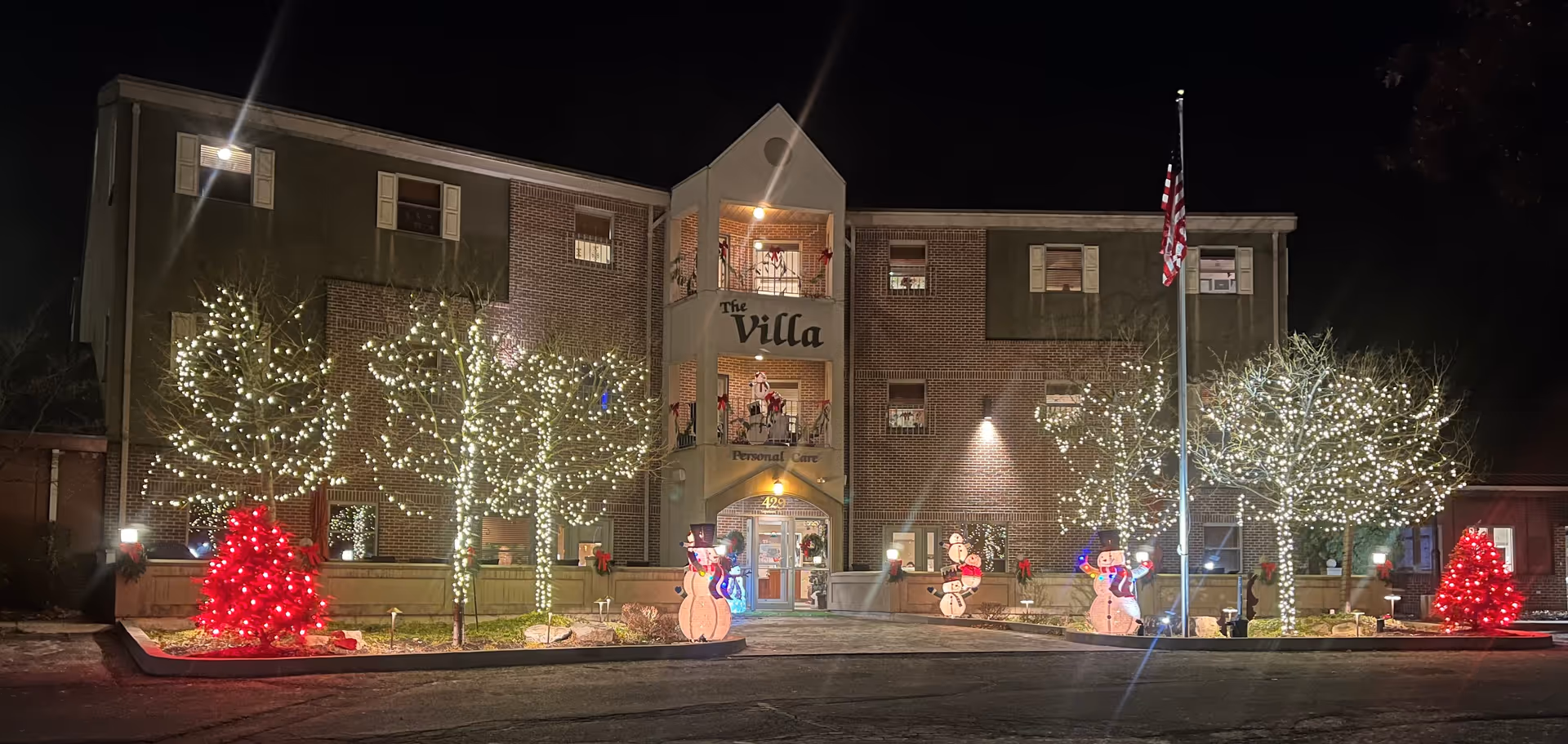 Front of a three-story brick assisted living building at night decorated with white string lights on trees, red lit bushes, and inflatable snowmen.