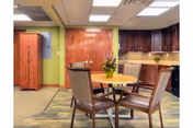 A cozy interior room featuring a round wooden table with four cushioned chairs around it. There is a flower arrangement on the table. The room has wooden cabinets along one wall, a green accent wall, and a carpeted floor with a patterned rug under the table.