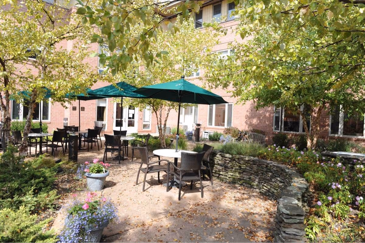 Outdoor patio area at The Villas at King David with round tables and chairs under green umbrellas, surrounded by trees and flowering plants, with a stone wall and a brick building in the background.