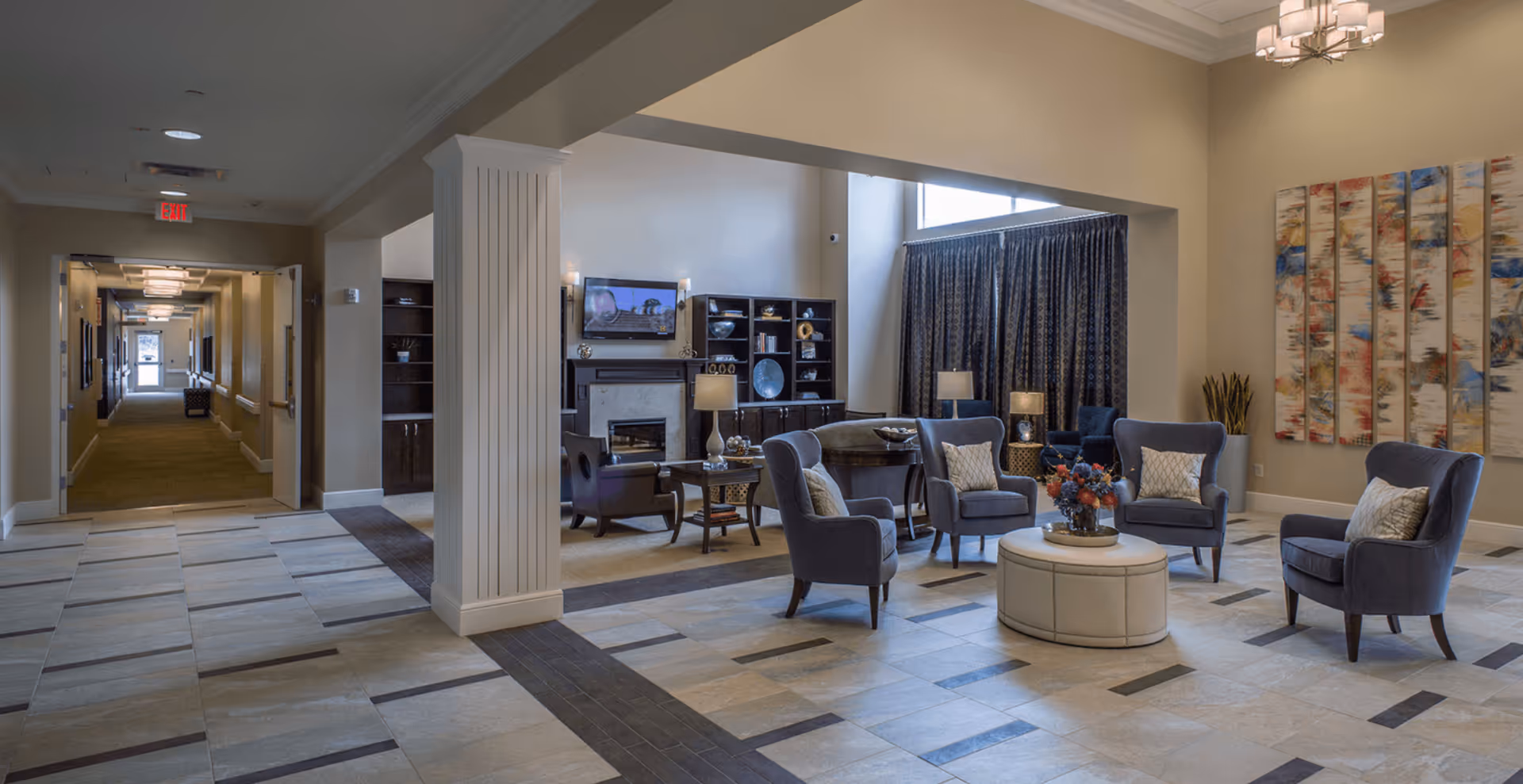 A spacious and well-lit common area in a senior living facility featuring several upholstered armchairs arranged around a round ottoman with a floral centerpiece. The room includes a fireplace with a mounted TV above it, built-in shelving, large windows with dark curtains, and colorful abstract artwork on the wall. A hallway extends from the seating area, leading to other parts of the facility.