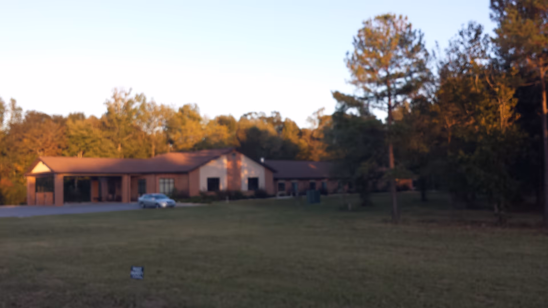 A single-story brick building with a covered entrance, surrounded by a large grassy area and trees under a clear sky during sunset.