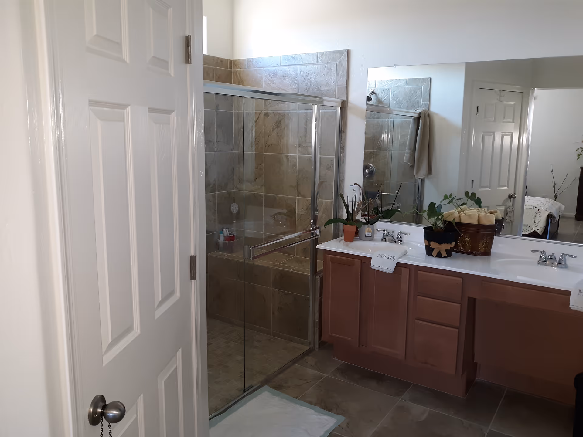 Bathroom with a glass-enclosed tiled shower, double-sink vanity with plants and towels, and an open door.