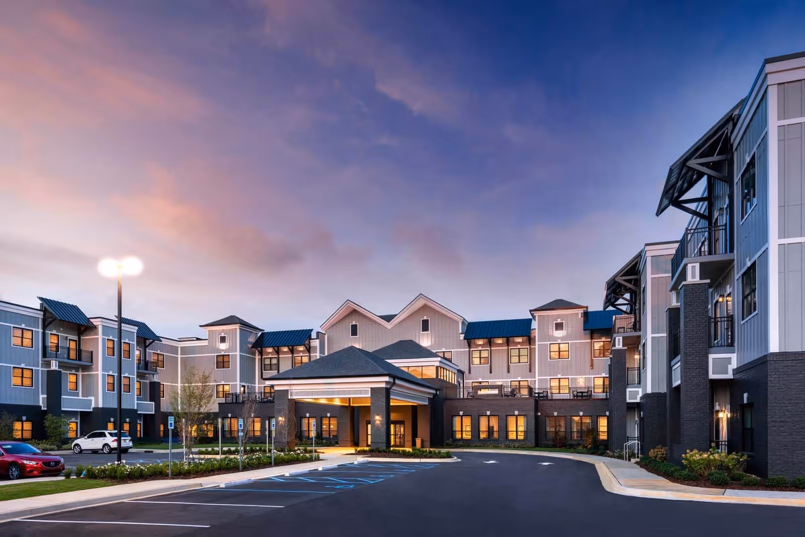 Exterior view of a modern senior living facility building at dusk with multiple floors, large windows, balconies, and a covered entrance. The parking lot in front has several cars and handicap parking spaces, with a streetlamp illuminating the area under a colorful sky.
