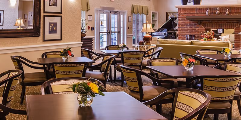 A cozy senior living facility common area with multiple tables and chairs arranged for dining or socializing. Each table has a small vase with flowers. The room features a large brick fireplace, a green sofa, a grand piano, and decorative lamps. There are framed pictures on the walls and a large mirror reflecting part of the room. Double glass doors lead outside.