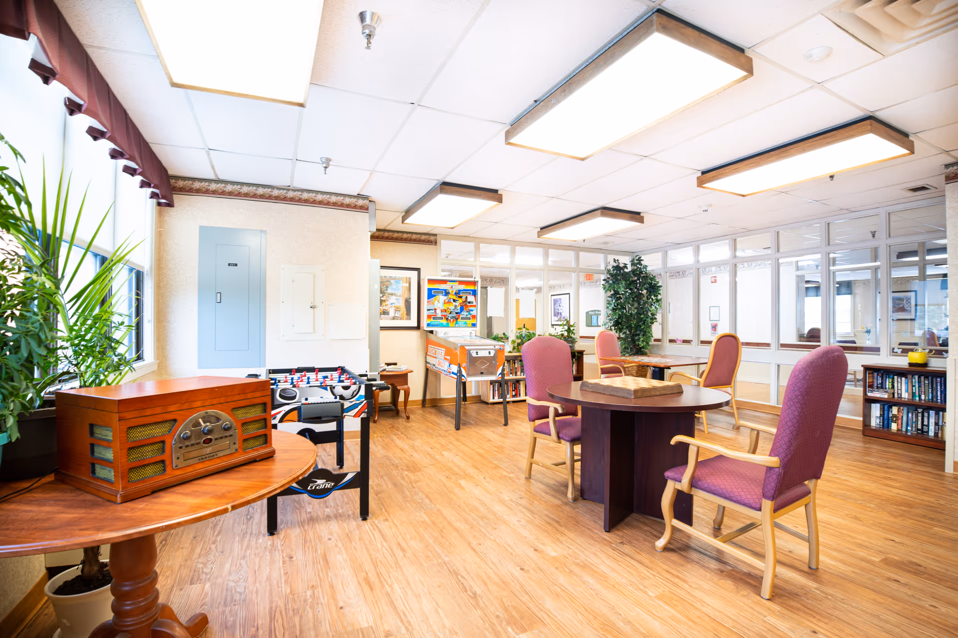 Bright communal activity room with a round table and upholstered chairs, foosball and arcade games, bookshelves, and large windows.