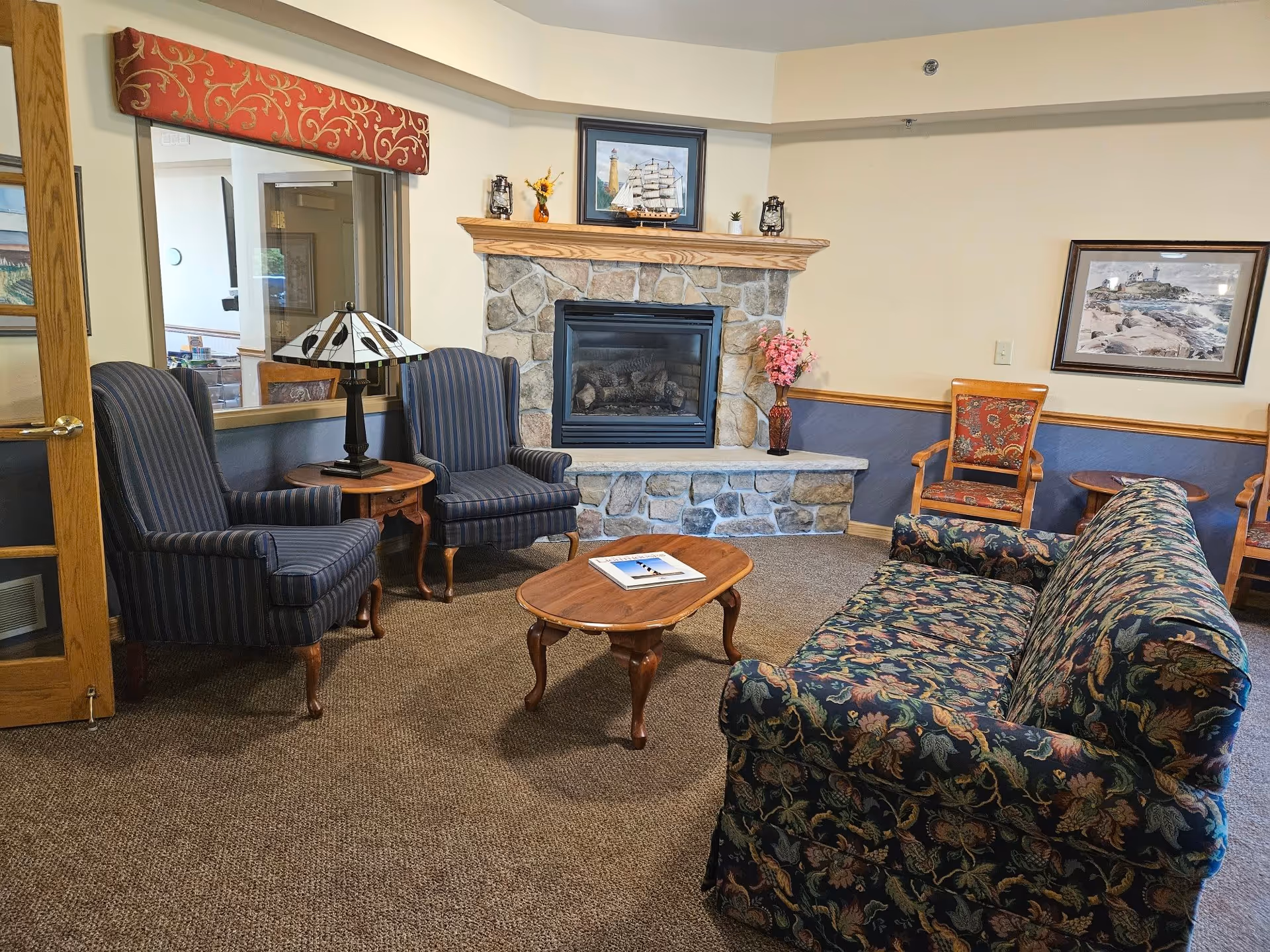 A cozy living room area with a stone fireplace, a floral patterned sofa, two striped armchairs, a wooden coffee table with a book on it, and a side table with a lamp. The walls are decorated with framed pictures and a vase with pink flowers is placed on the fireplace hearth.