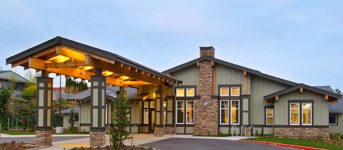 Exterior view of The Arbor at Bremerton senior living facility showing a single-story building with a covered entrance supported by pillars, stone accents, multiple windows, and a driveway in front.