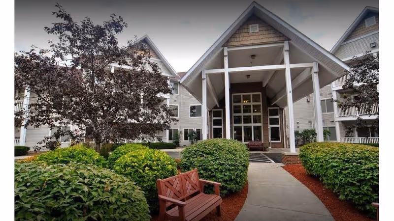 Entrance of Charter Senior Living of Kenosha featuring a covered porch with white pillars, surrounded by well-maintained bushes and trees, with a wooden bench on the left side of the walkway.