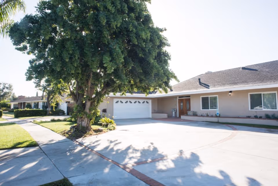 Exterior view of a single-story residential building with a large tree in the front yard, a wide driveway, and a garage with a white door. The building has beige walls, a dark shingled roof, and two front doors with glass panels. There is a sidewalk and green lawn surrounding the property.