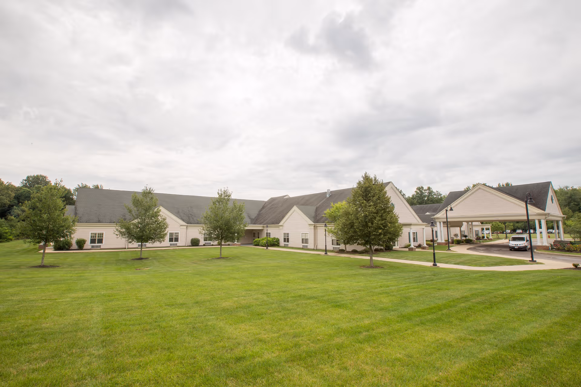 Exterior view of Riverview Pointe Care Center showing a large, single-story building with a covered entrance driveway, surrounded by a well-maintained lawn and several trees under a cloudy sky.