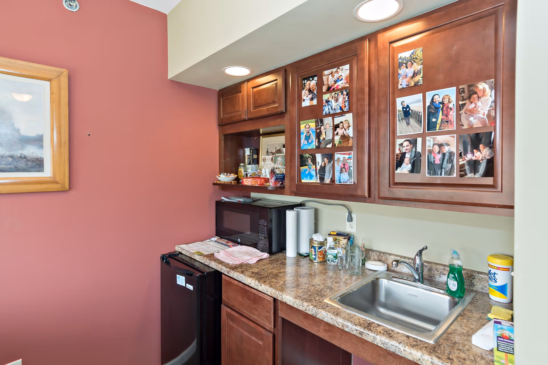 A small kitchen area with a countertop, stainless steel sink, microwave, mini refrigerator, and wooden cabinets. The cabinets have several family photos attached to them. Various kitchen items such as paper towels, dish soap, and cleaning wipes are on the counter. The wall is painted in two colors, beige and reddish-brown, with a framed painting hanging on the reddish-brown wall.
