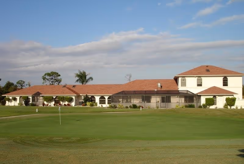 A single-story building with a red-tiled roof and white walls, surrounded by a well-maintained green lawn and a golf putting green in the foreground under a partly cloudy sky.