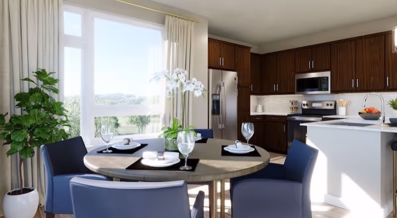 A bright kitchen and dining area with a round table set for four, featuring plates, napkins, and wine glasses. The kitchen has dark wooden cabinets, stainless steel appliances including a refrigerator, stove, and microwave. There is a large window with curtains letting in natural light, and a potted plant and white orchids add greenery to the space.