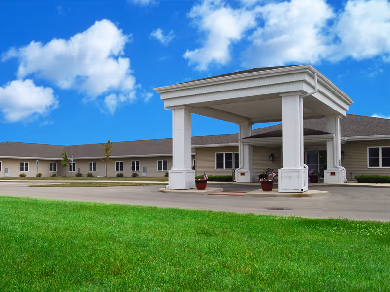 Exterior view of a single-story senior living facility building with a covered entrance driveway, green lawn in the foreground, and a bright blue sky with scattered clouds.