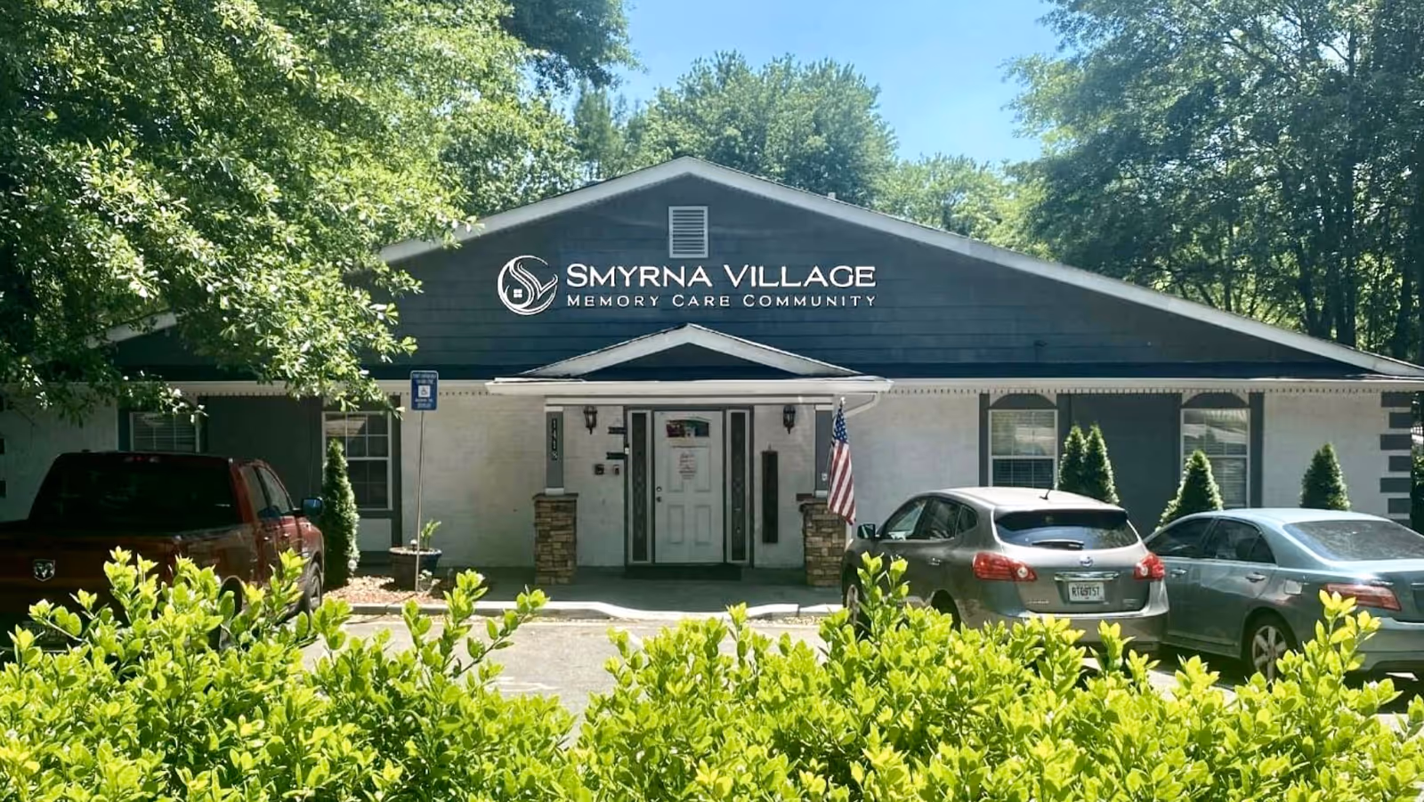 Front exterior view of Smyrna Village Memory Care Community building with a peaked roof, entrance door with small porch, an American flag, several parked cars, and green bushes and trees surrounding the parking area.