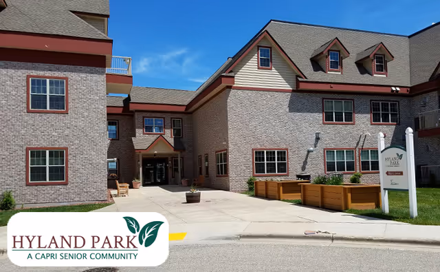 Exterior view of Hyland Park, a senior community building with brick walls, multiple windows, and a sloped roof under a clear blue sky. There is a paved walkway leading to the entrance, with planters and a sign displaying the community name.