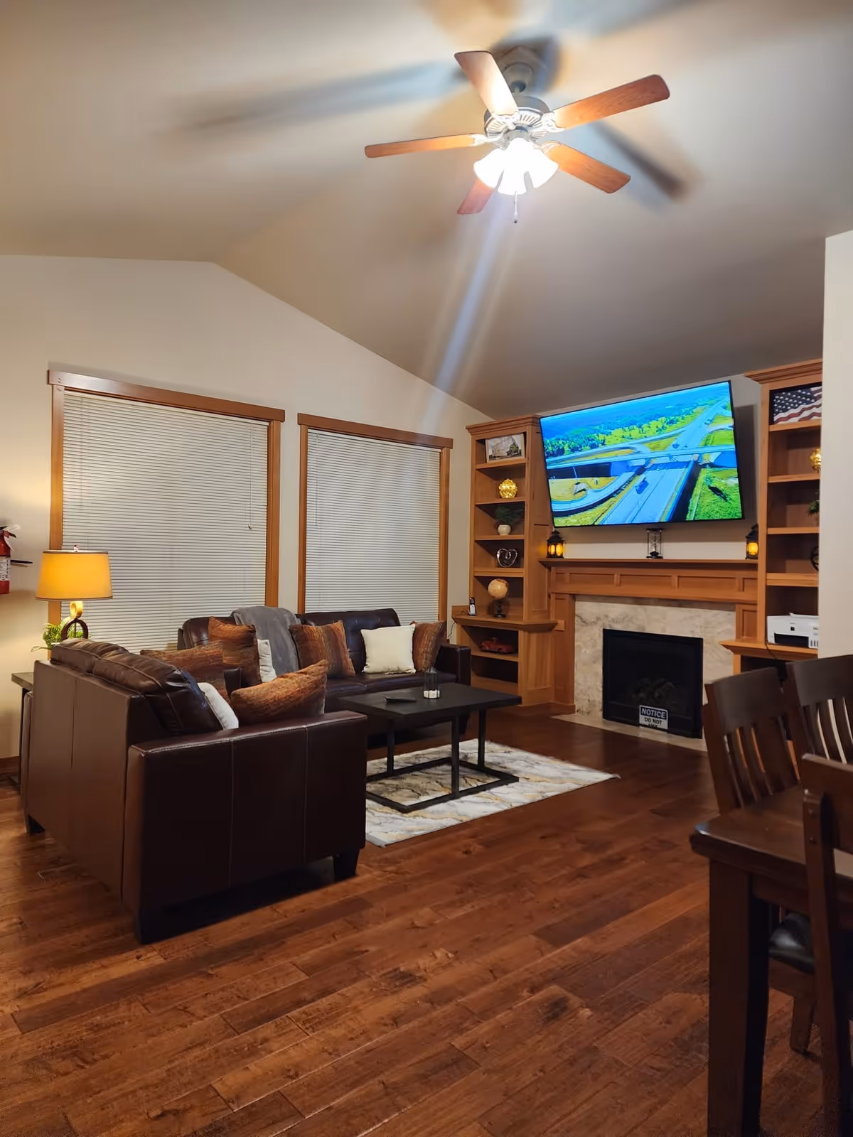 A cozy living room with wooden floors, a ceiling fan with lights, two large windows with blinds, a brown leather sectional sofa with multiple cushions, a black coffee table on a patterned rug, a wooden built-in shelving unit surrounding a fireplace, and a large flat-screen TV mounted above the fireplace displaying an aerial view of a highway interchange.