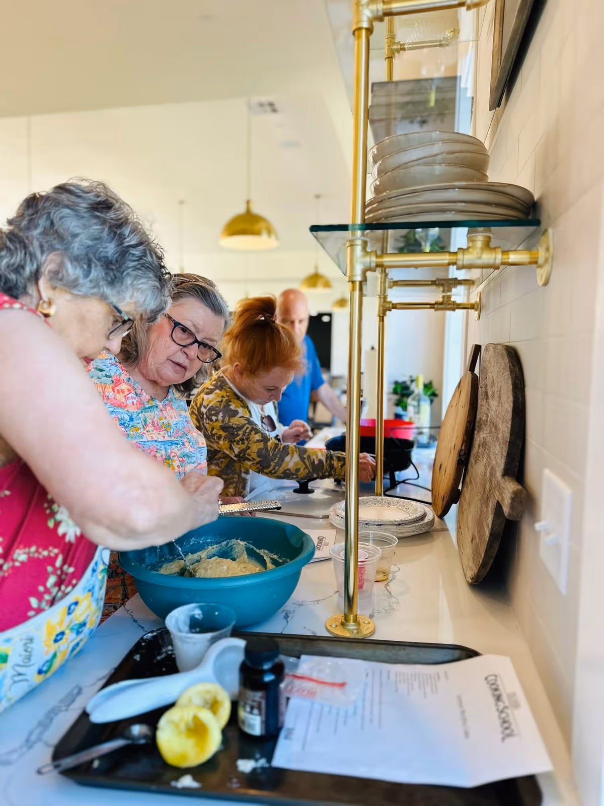 Four elderly people standing in a kitchen preparing food together. One woman in the foreground is mixing batter in a large blue bowl, while others are focused on their tasks along the counter. The kitchen has white countertops, brass pipe shelving with stacked plates, and wooden cutting boards on the wall.